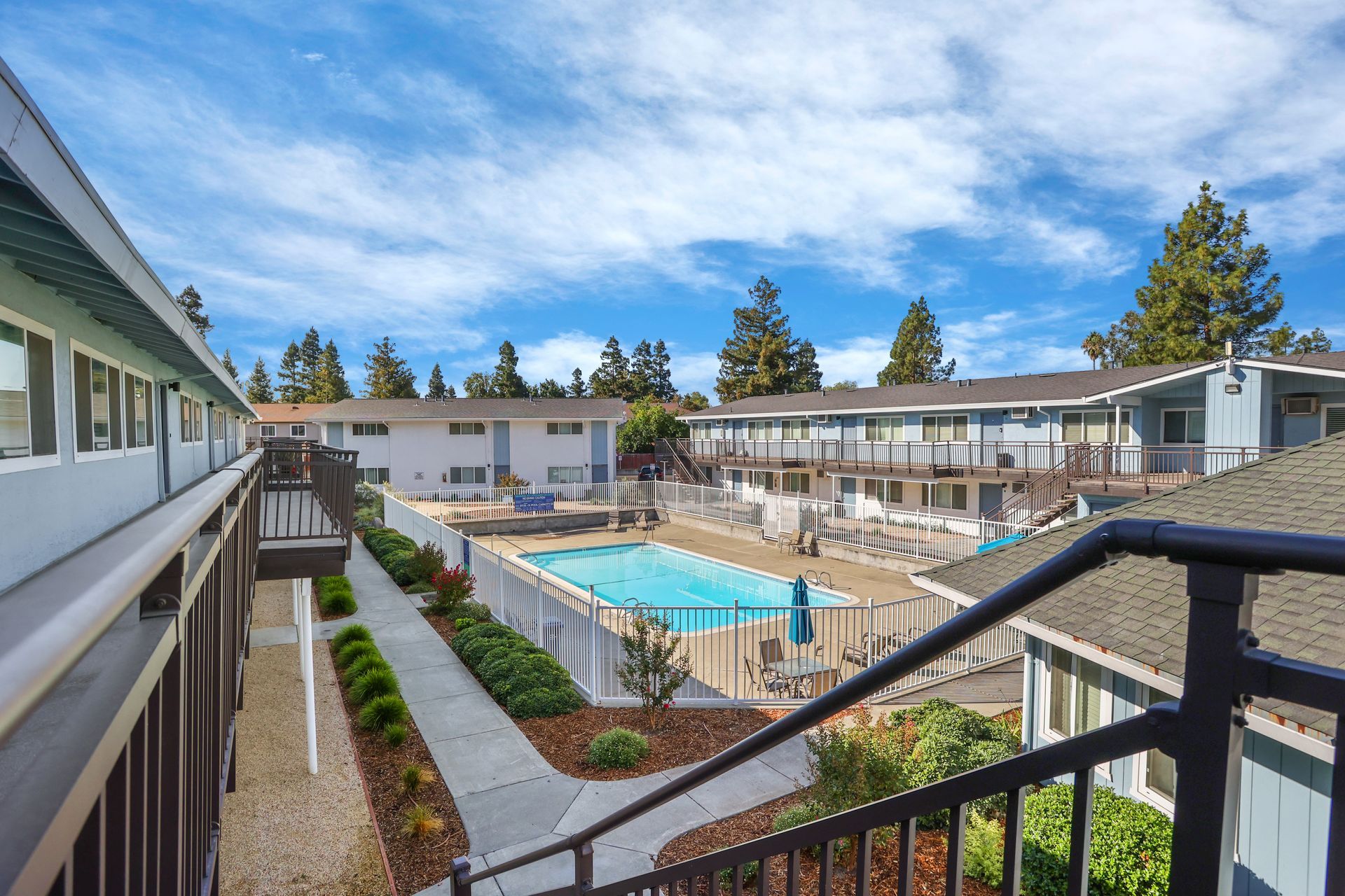 Apartment courtyard with a blue swimming pool, patio seating, and stair railings on a sunny day