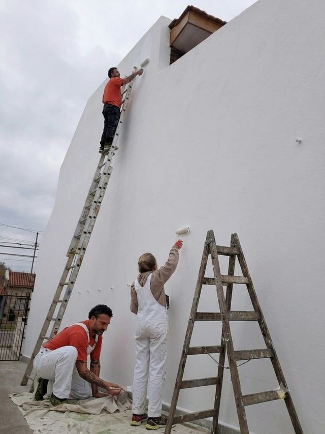 Trabajadores pintando una pared blanca alta usando escaleras y rodillos al aire libre