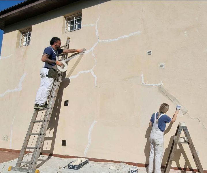 Dos trabajadores reparan grietas en una pared exterior de color beige mientras están subidos a escaleras.