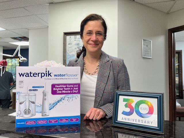 A person smiles behind a counter with a Waterpik box and a 30th-anniversary framed sign in a dental office setting.