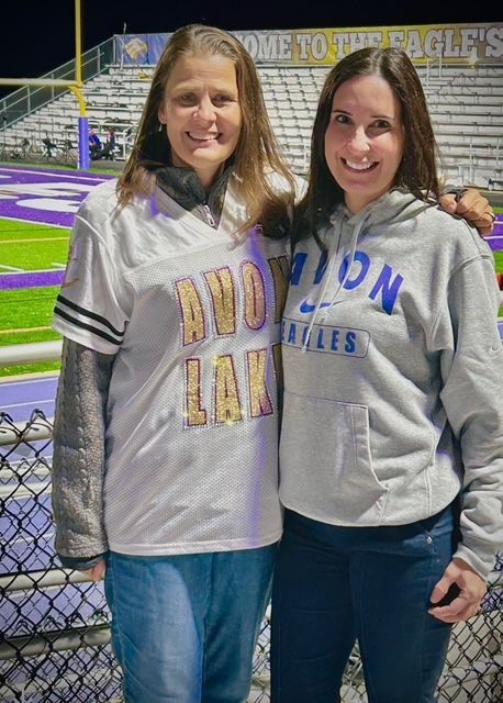 Two smiling women standing arm-in-arm on a high school football field at night, wearing Avon Lake spirit wear.