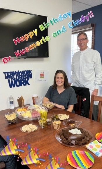 A person seated at a table with snacks and a chocolate cake next to a standing colleague in an office with party decor.