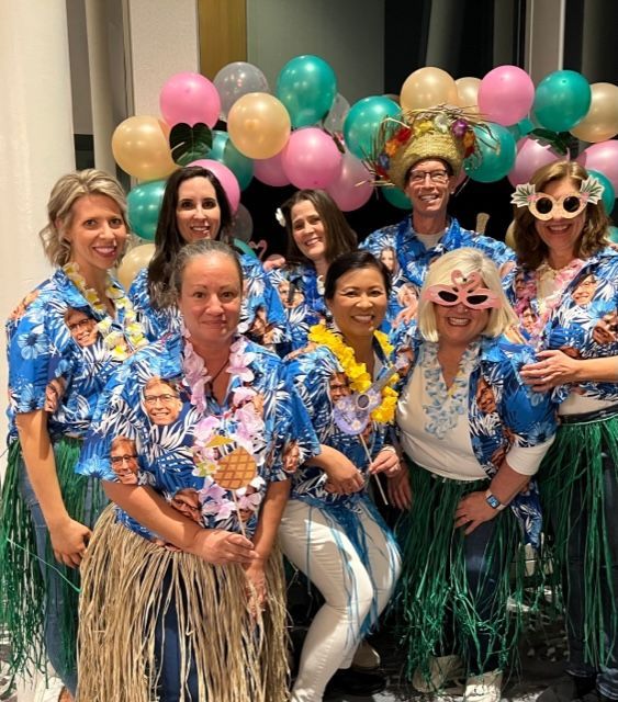 A group poses at a party wearing matching floral shirts and grass skirts, surrounded by colorful balloons and leis.