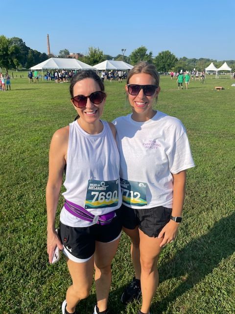 Two people wearing sunglasses and race bibs stand in a grassy field in front of event tents on a sunny day.