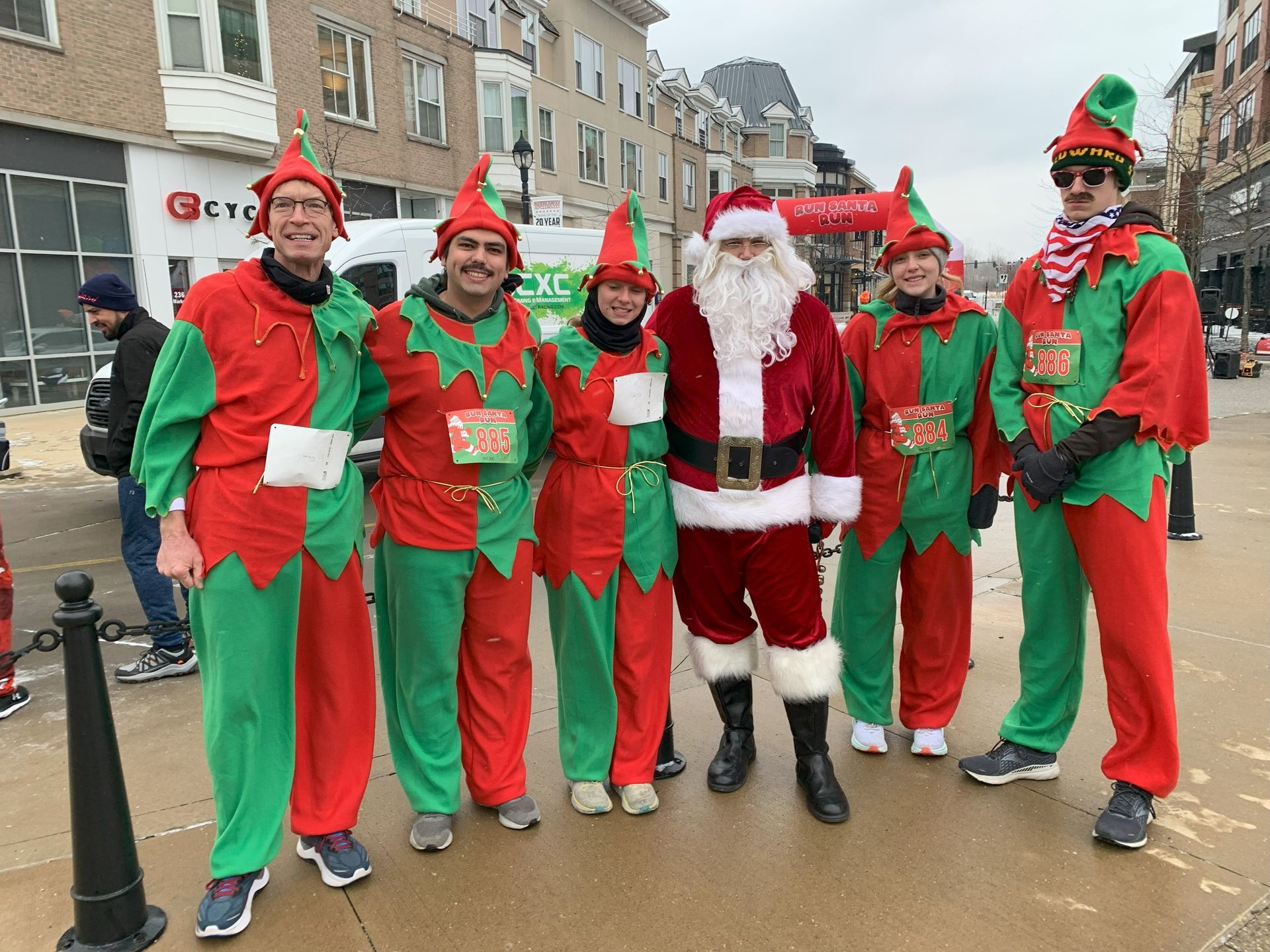 Santa Claus stands in the center surrounded by five people in green and red elf costumes on an urban street.