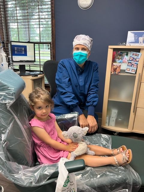 A healthcare worker wearing scrubs and a mask sits behind a young child holding a stuffed animal in a dental chair.