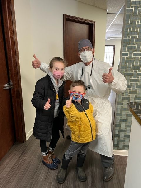 A healthcare professional in protective gear and two children in winter clothes pose giving a thumbs-up in a dental office.