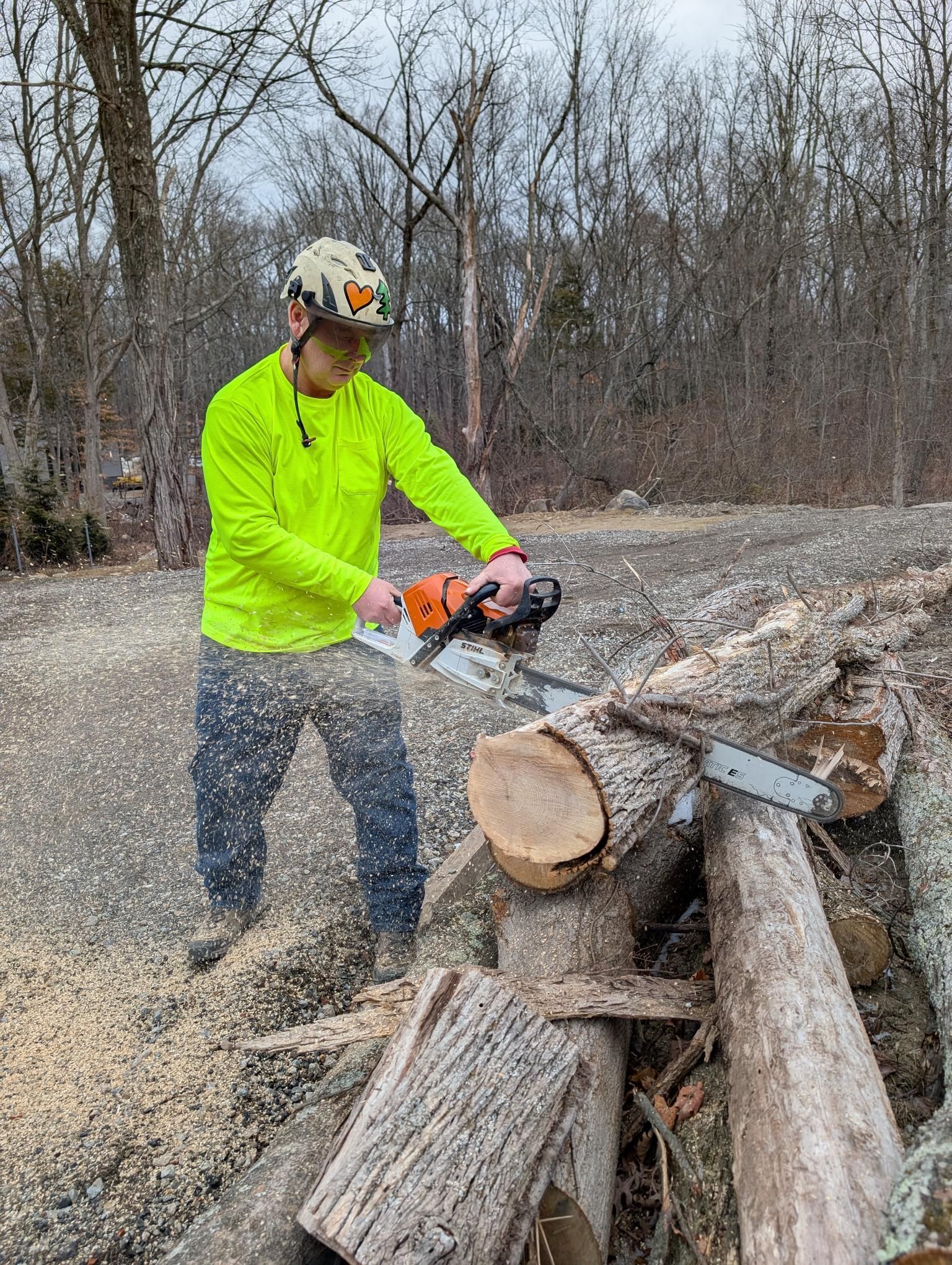 Worker in neon shirt cutting a large log with a chainsaw outdoors near a woodpile