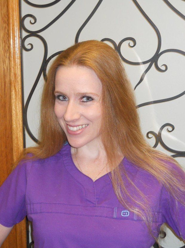Smiling woman with long red hair in a purple top, standing before a decorative metal wall