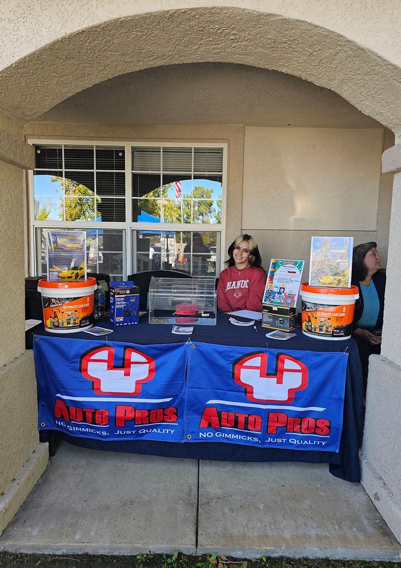 A person sits behind an Auto Plus branded table with promotional buckets and materials under a stucco archway.