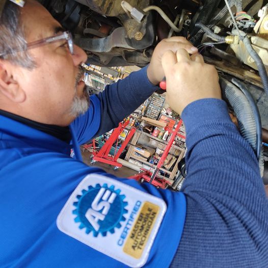 Technician in blue uniform inspecting machinery inside a tight engine compartment | Auto Pros