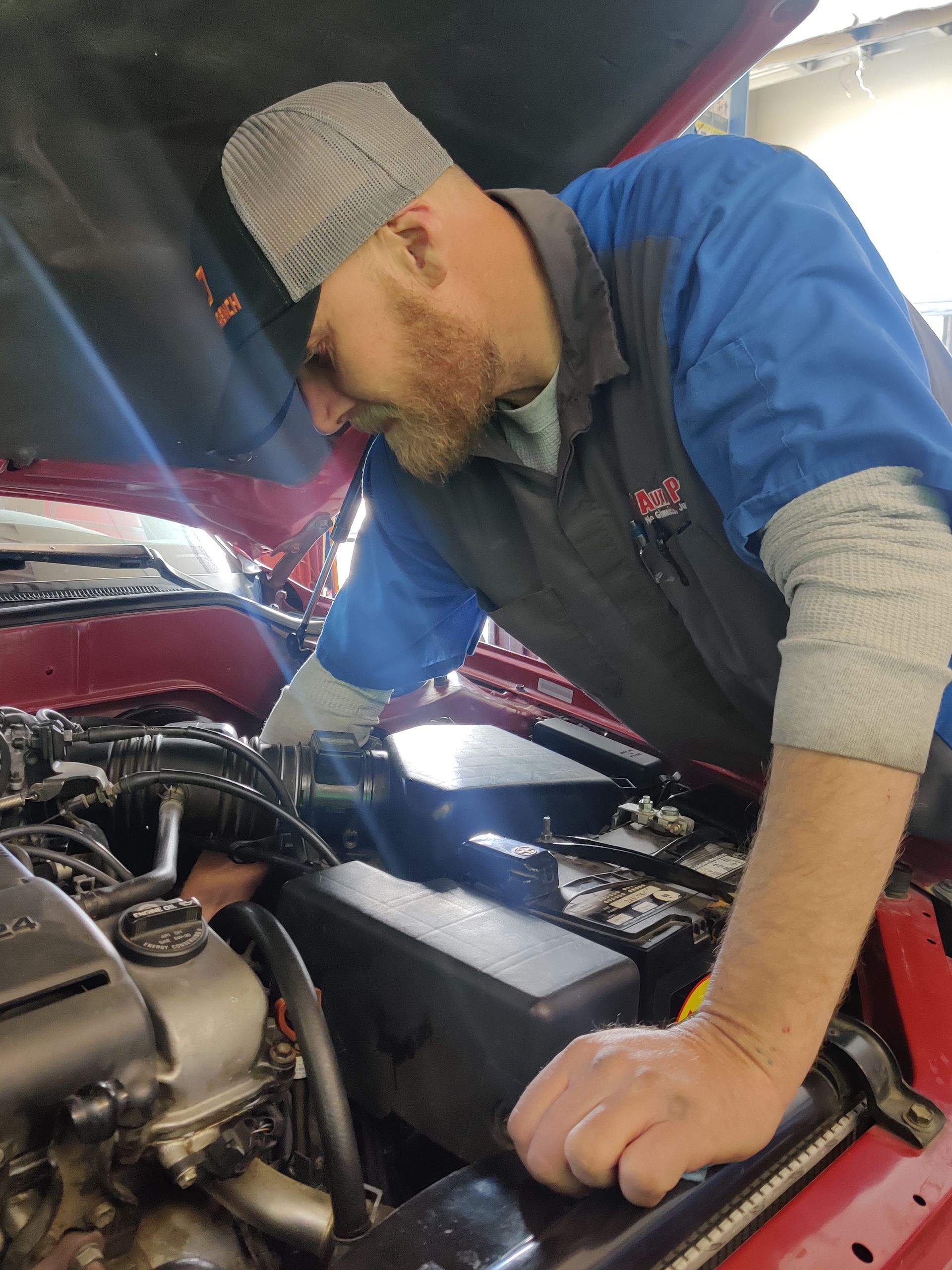 Mechanic in blue shirt inspecting engine under an open car hood | Auto Pros