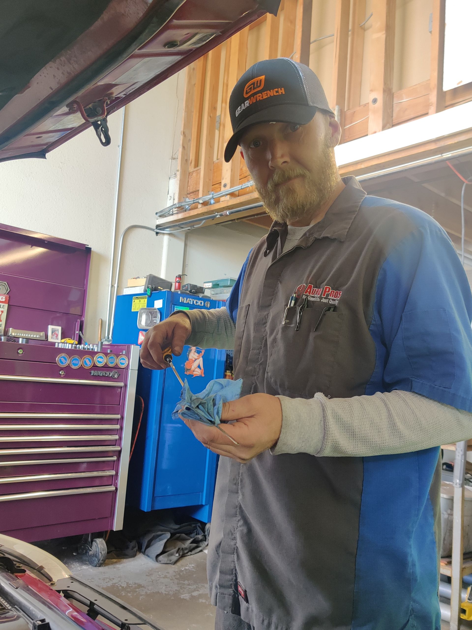 Man in gray work shirt holding a blue part in a garage workshop beside tool cabinets | Auto Pros