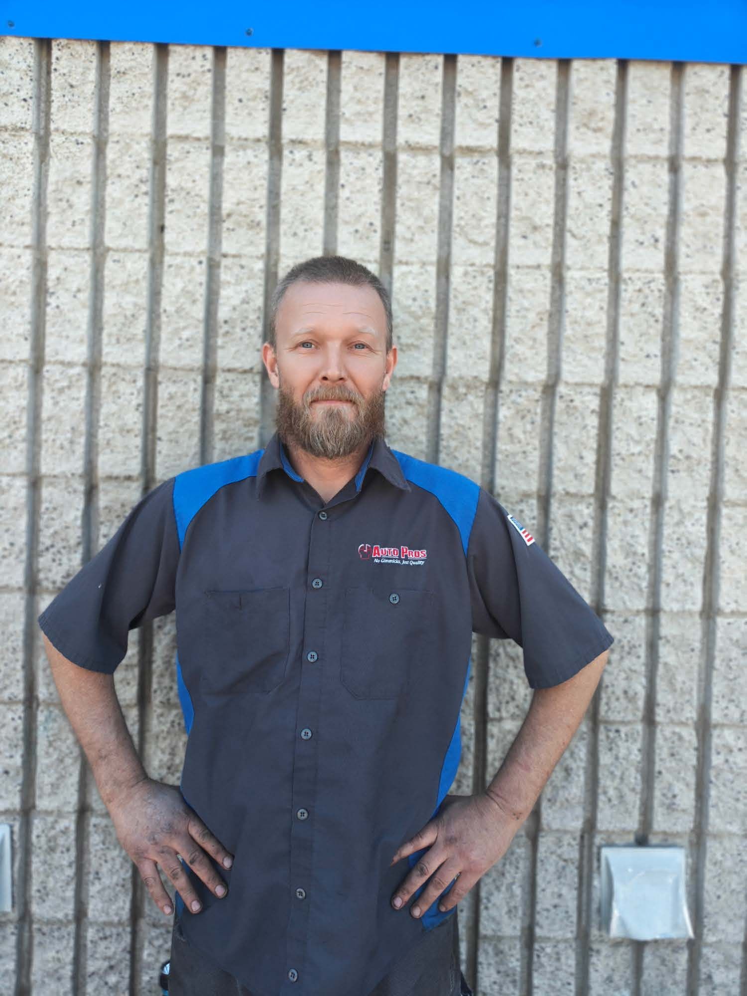 Man standing with hands on hips in front of a concrete wall, wearing a dark shirt with blue shoulders. | Auto Pros