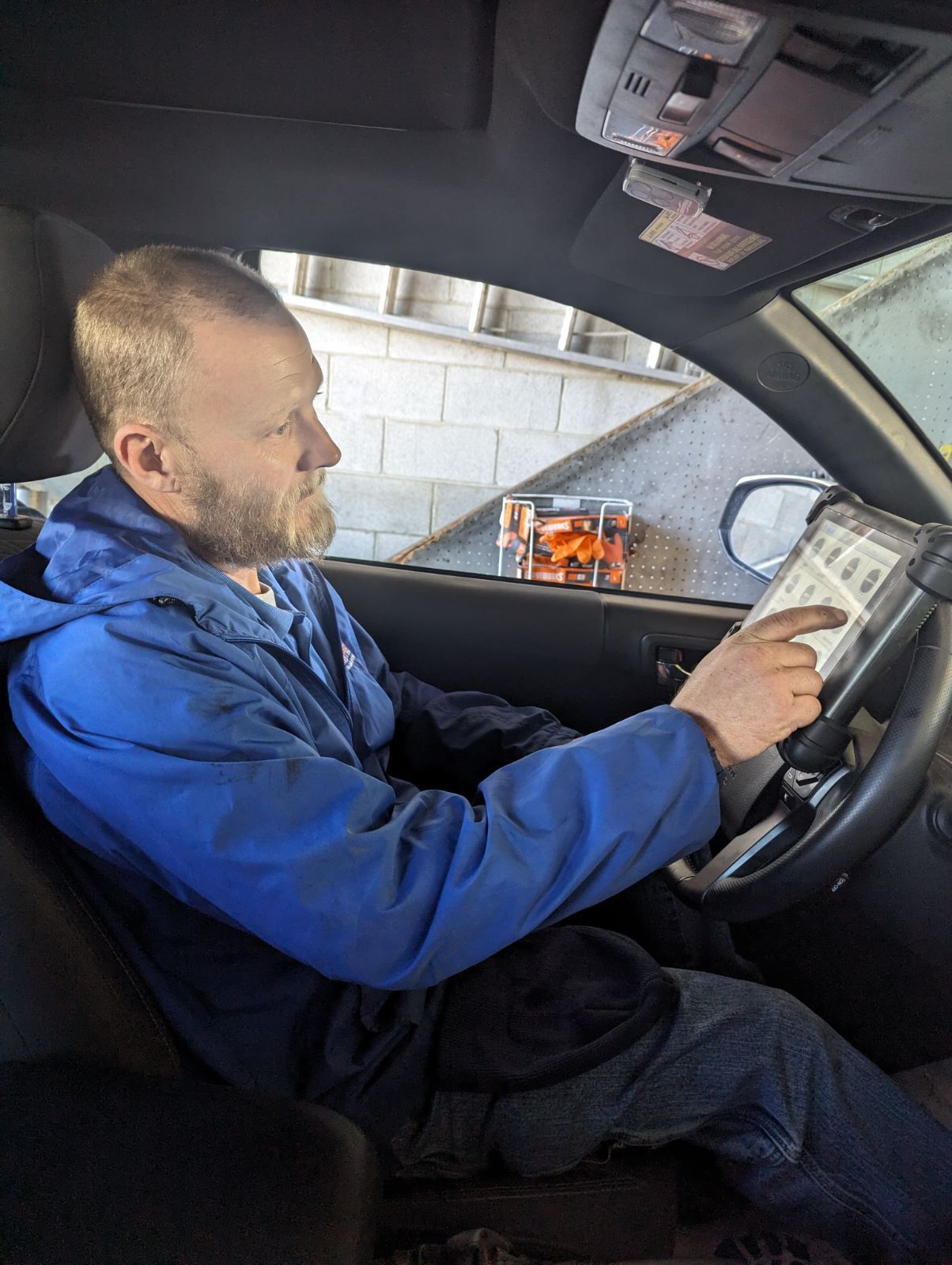 Man in a blue jacket sitting in a car, reading a paper while parked indoors. | Auto Pros