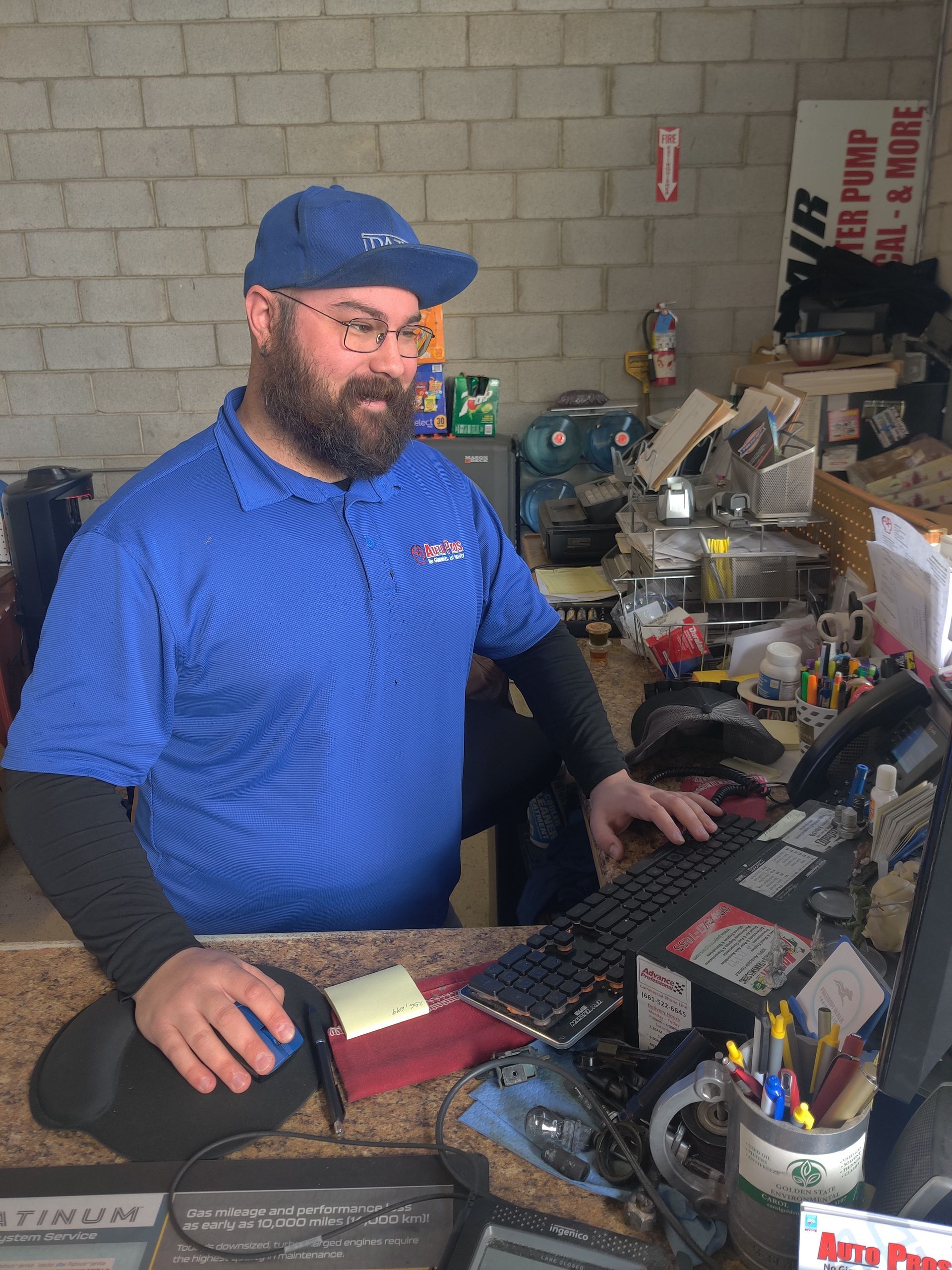 Man in blue shirt and cap sitting at a cluttered desk with a laptop and office supplies | Auto Pros