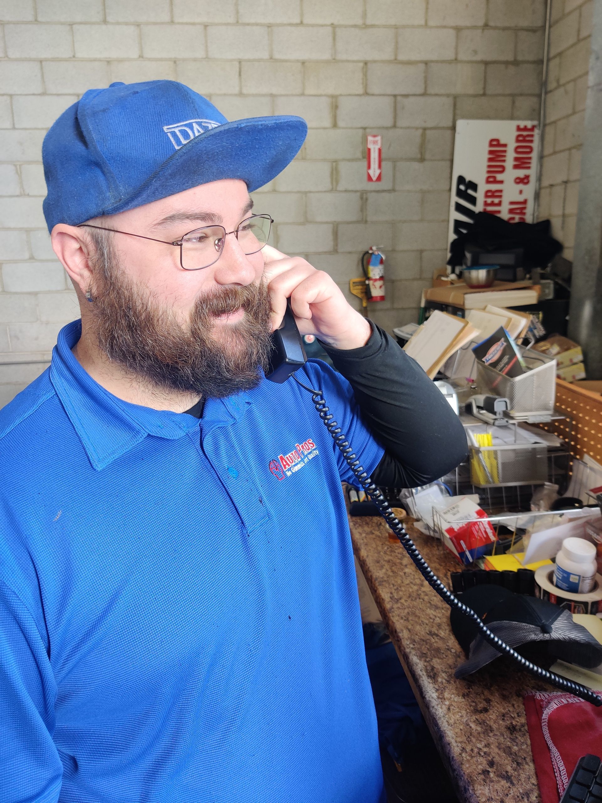Smiling bearded man in blue cap and shirt talking on a phone beside a cluttered workbench | Auto Pros