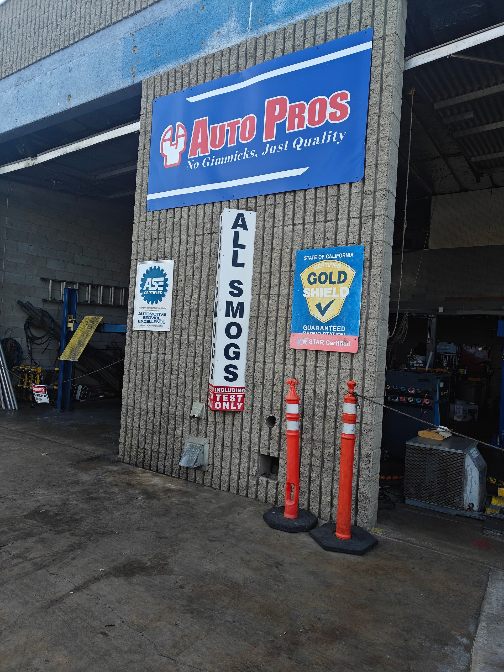 Auto repair shop entrance with blue sign, hanging “All Smogs” banner, and orange cones by the doorway | Auto Pros