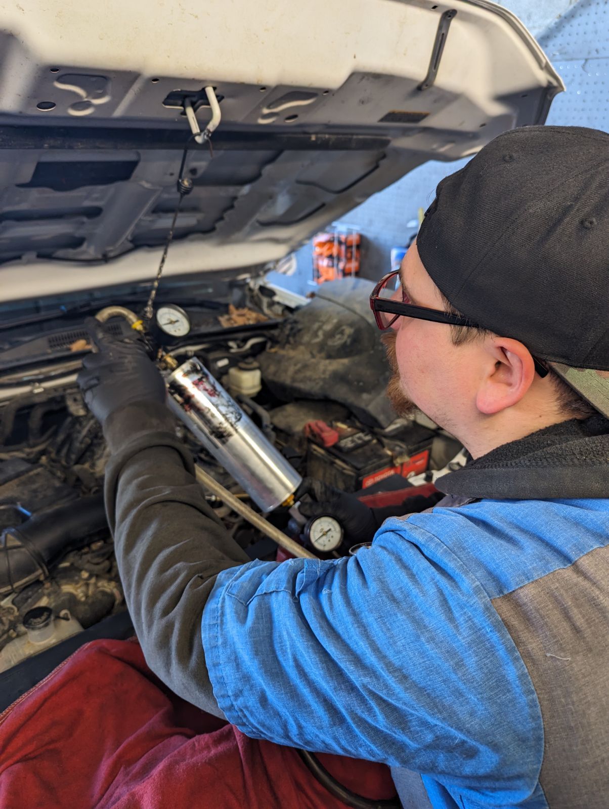 Mechanic in blue jacket and black cap working under a car hood with a tool near the engine | Auto Pros
