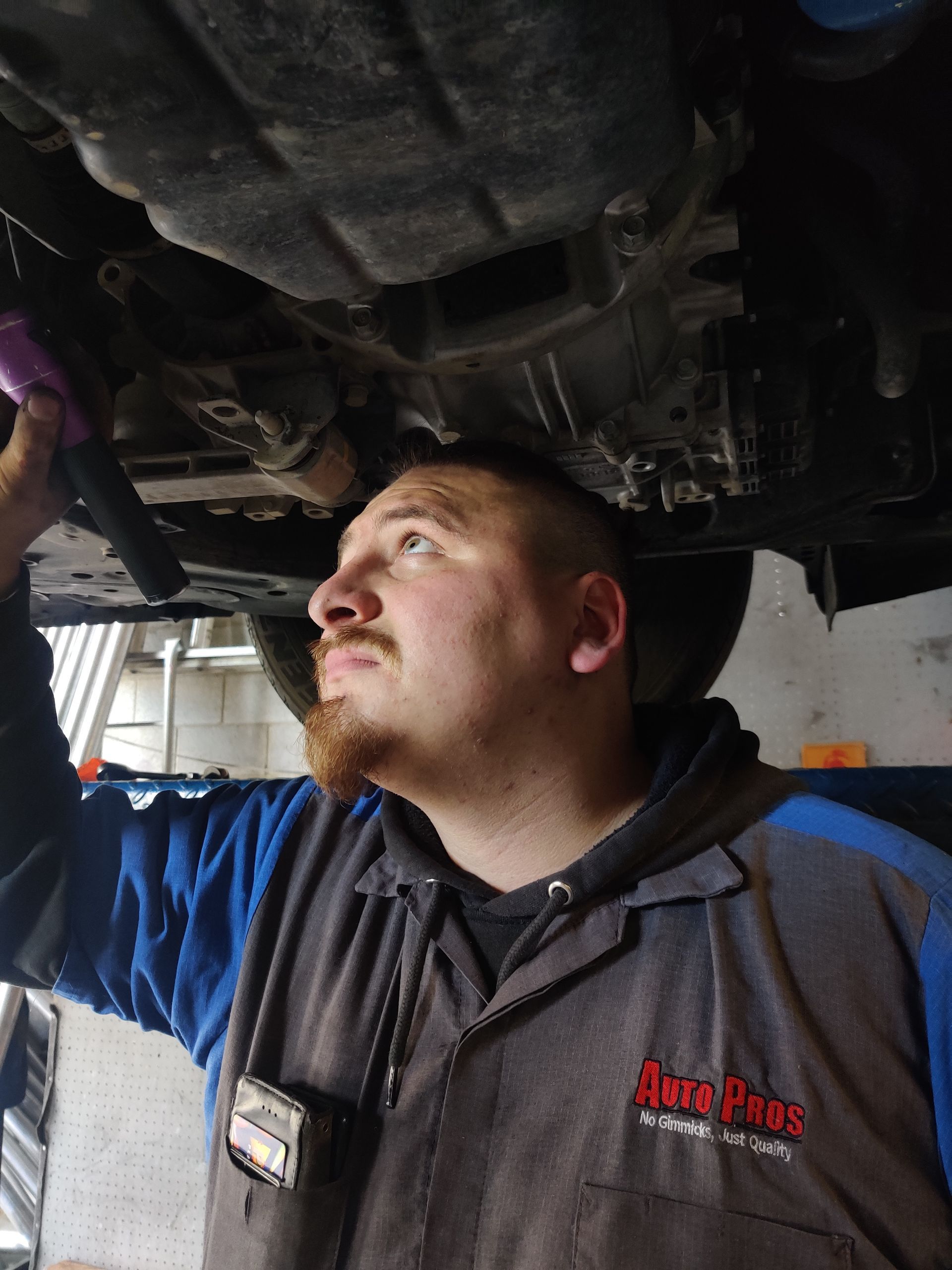 Mechanic in blue coveralls inspecting the underside of a vehicle in a garage | Auto Pros