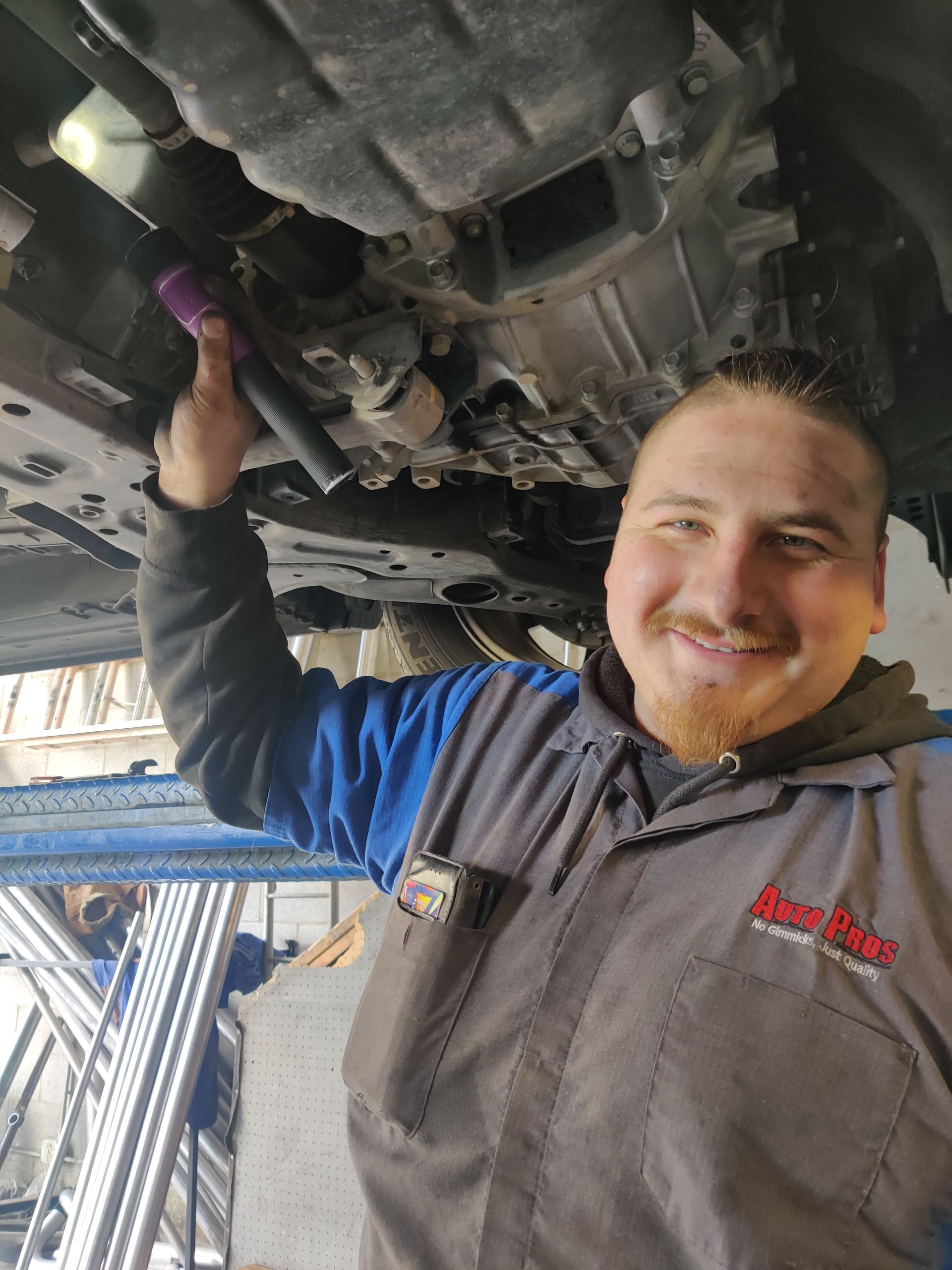 Mechanic smiling under a vehicle in a garage, holding tools while working beneath the chassis | Auto Pros