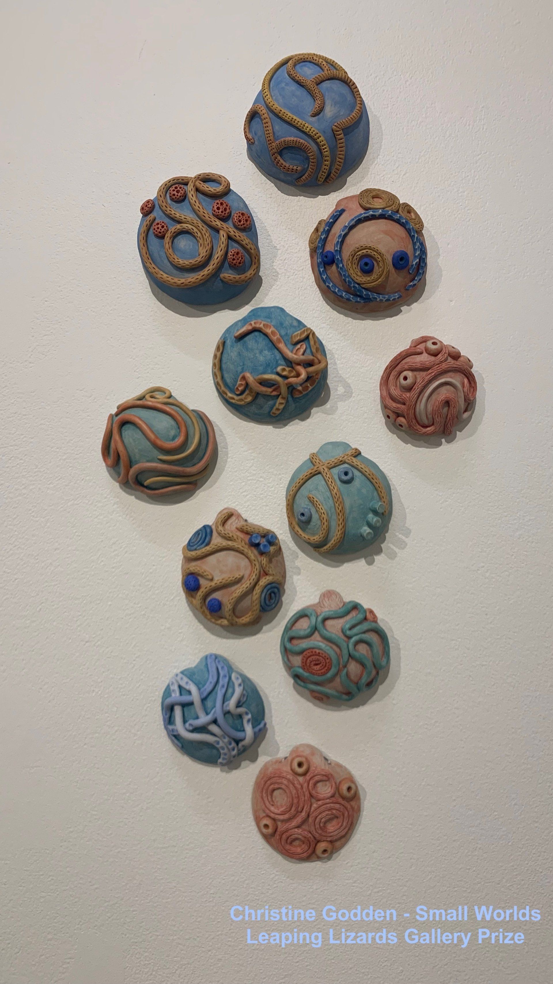 Ten textured, spherical clay sculptures with varied swirling patterns and earthy, blue, and terracotta tones on a white wall.