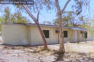 A pale, single-story rectangular building, identified as an art shed in 1990, set among trees on a dry, dirt lot.