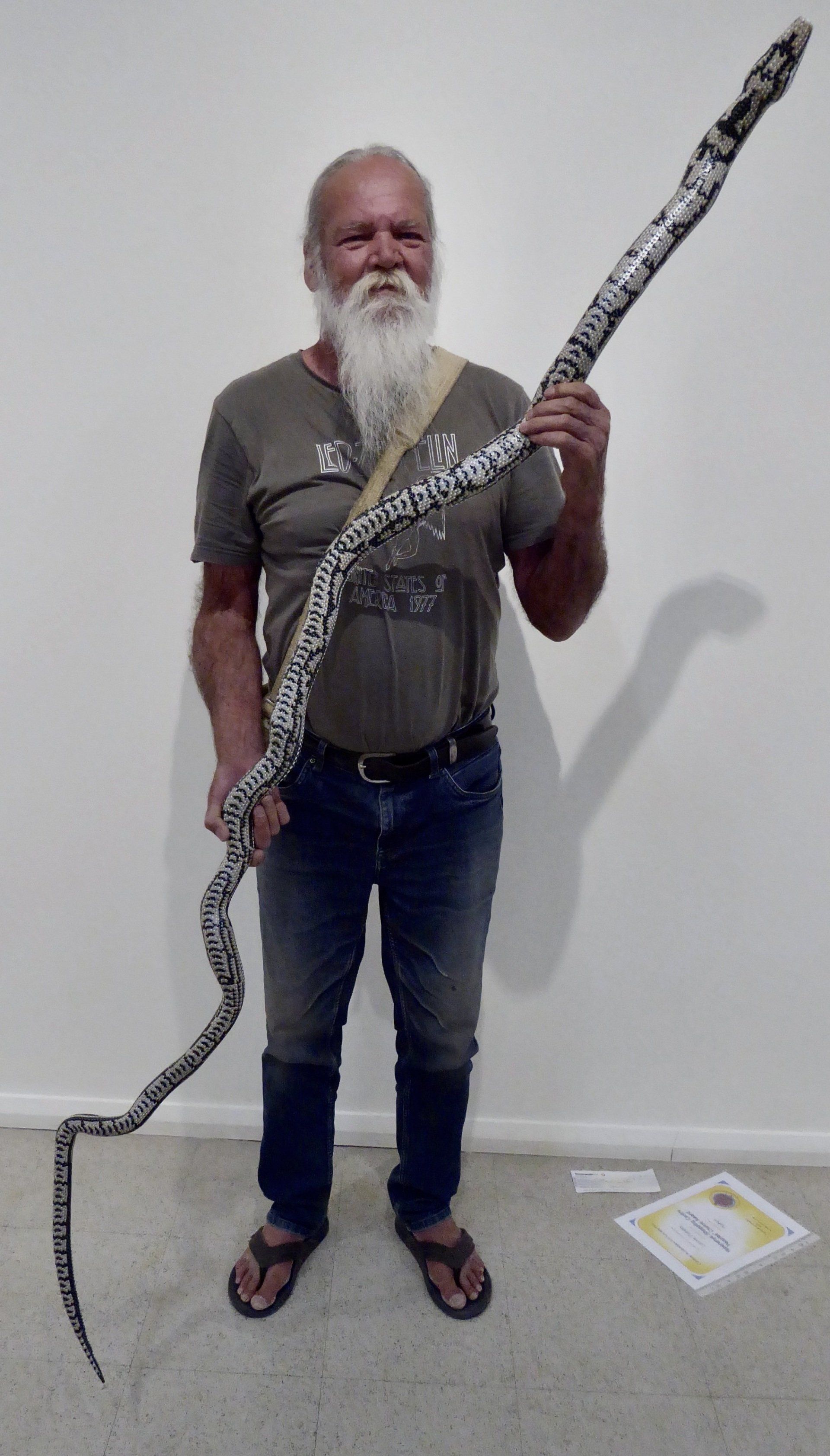A person with a long white beard holding a large, patterned snake sculpture against a plain white wall.
