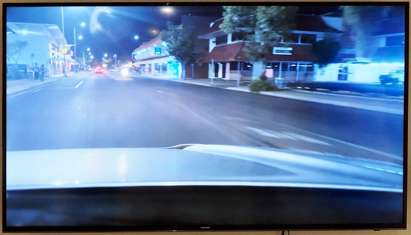 View from a car windshield at night, driving along a street lined with buildings and streetlights.