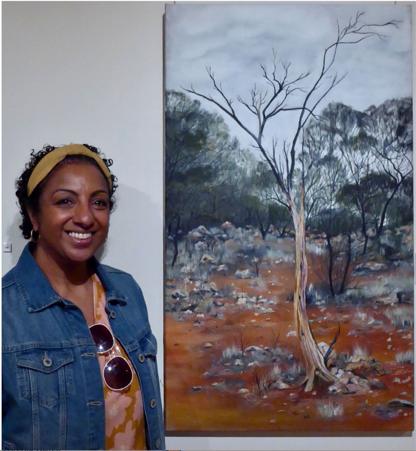 A person smiling next to a tall oil painting of a bare tree in an arid, red-earth landscape under a cloudy sky.