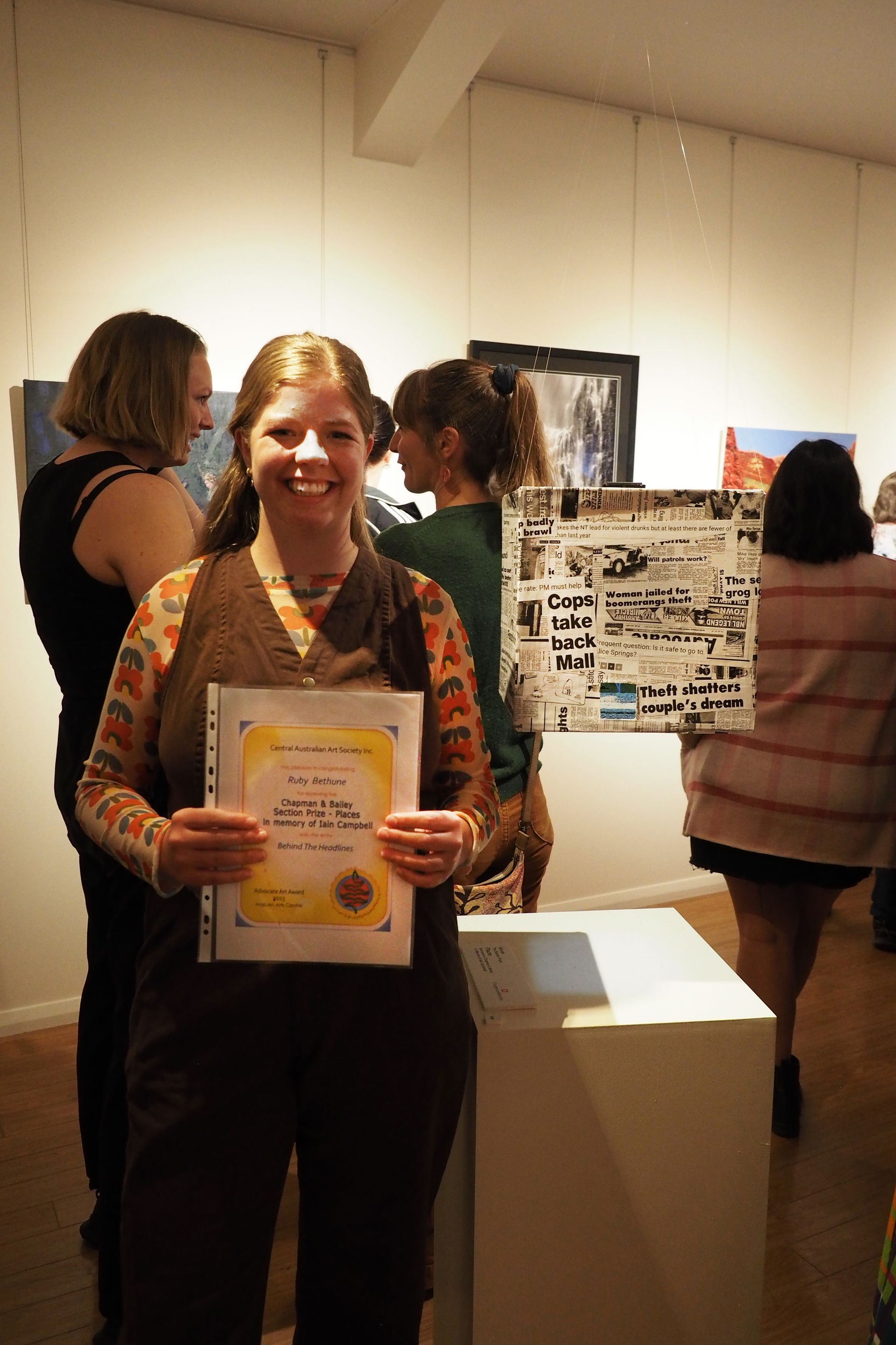 A smiling person holds a certificate at an indoor art gallery, with other people and artwork in the background.