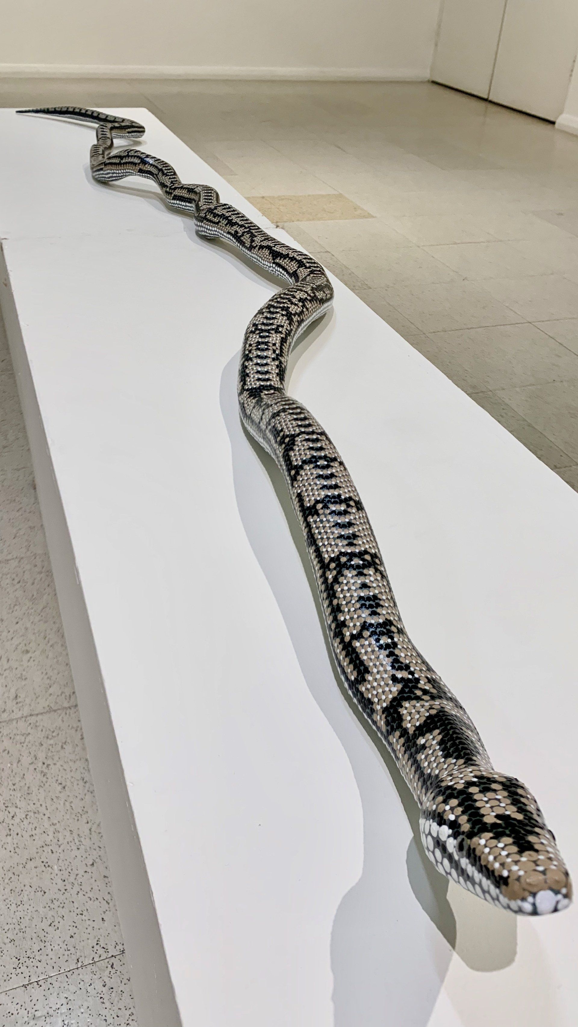A long, intricately textured sculpture of a snake with patterned scales rests on a white gallery pedestal.