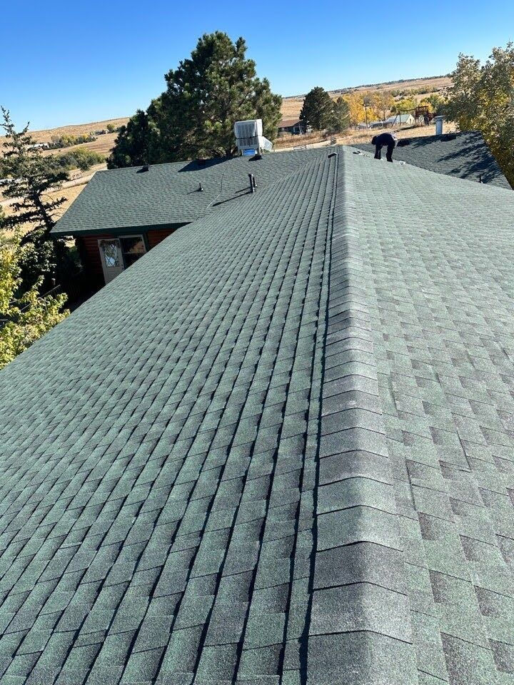 Wide view of a shingled roof with two workers on top, surrounded by trees and dry hills under a clear sky