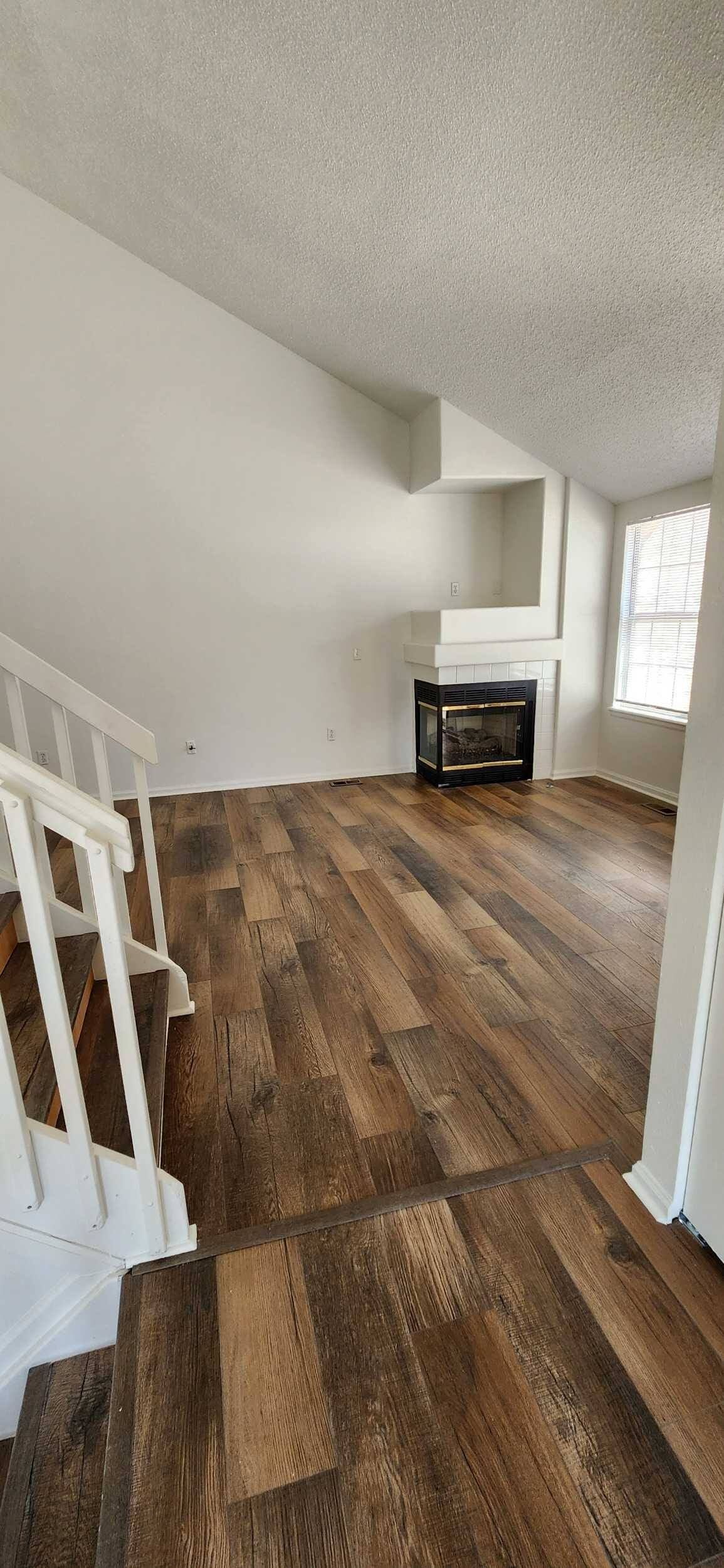Empty living room with wood flooring, staircase, and a fireplace under a slanted ceiling.