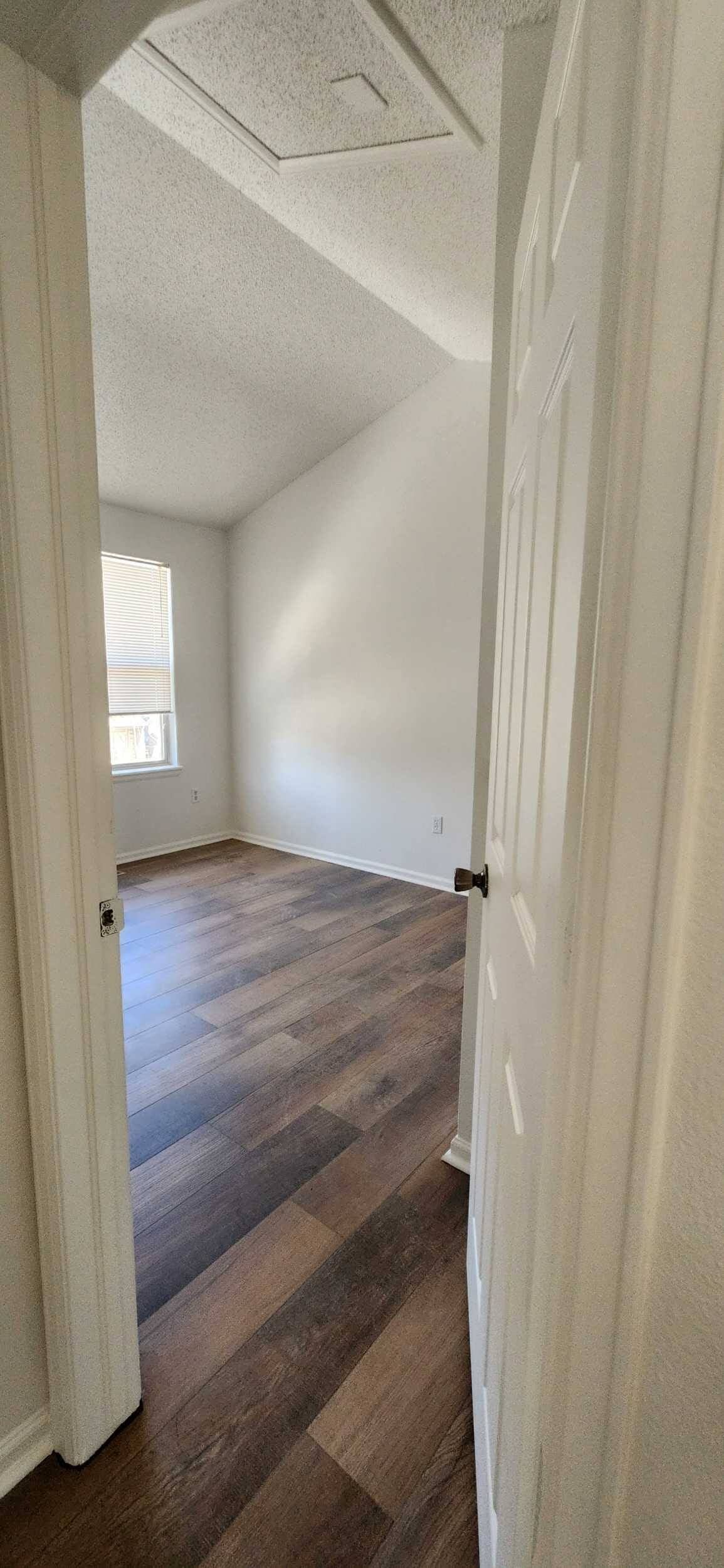 Doorway view into an empty room with dark wood floors, white walls, and a vaulted ceiling.