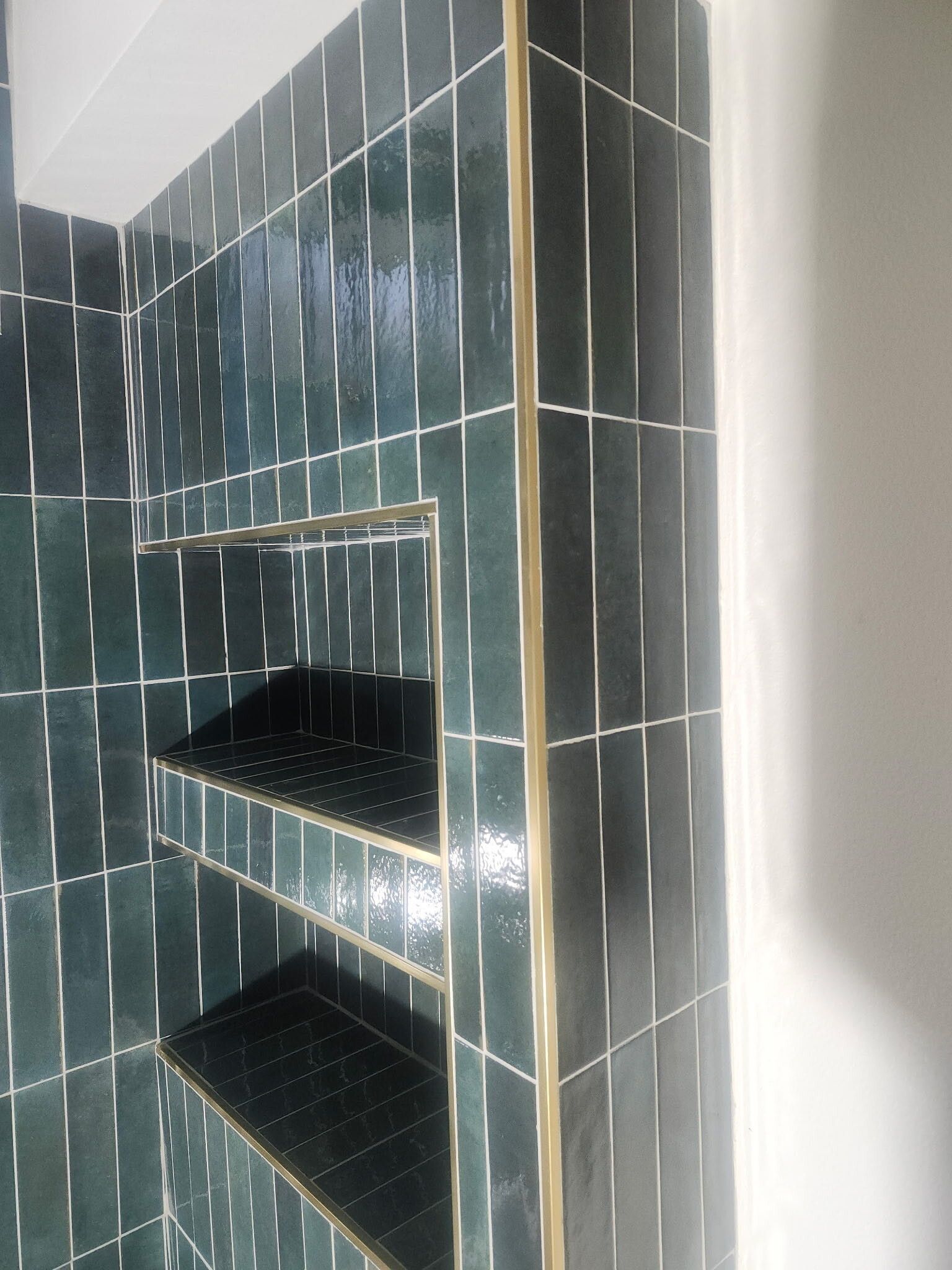 Dark green tiled shower niche with two shelves and white grout on a bathroom wall
