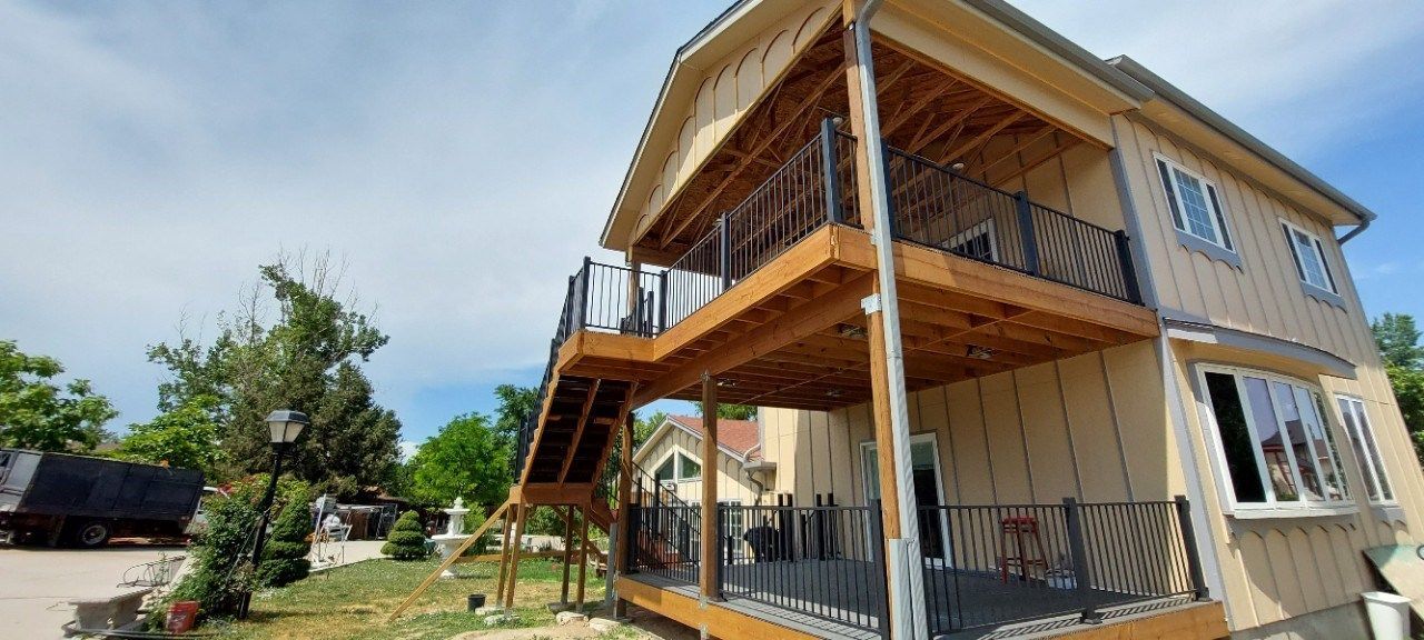Two-story cream house with wood balconies, external stairs, and a small yard on a sunny day