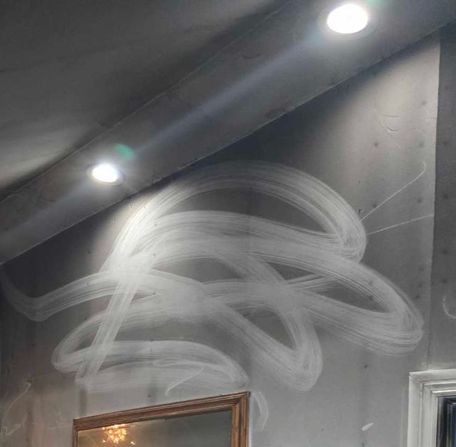 White spray-paint graffiti on a gray indoor wall beneath two ceiling lights.