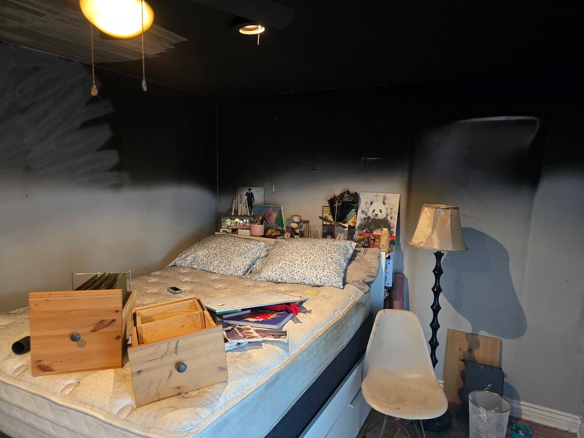 Cozy bedroom with a bed, nightstand, lamp, chair, and storage boxes under dark walls.
