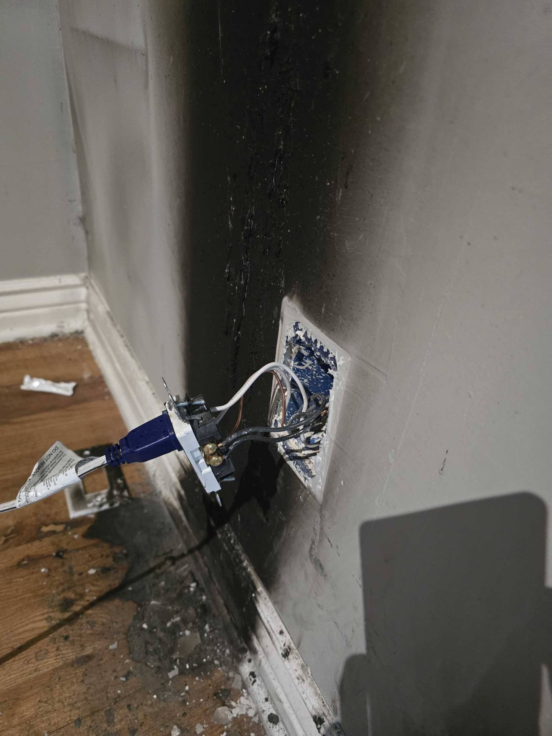 Damaged wall outlet with exposed wires and black scorch marks near the floor and baseboard
