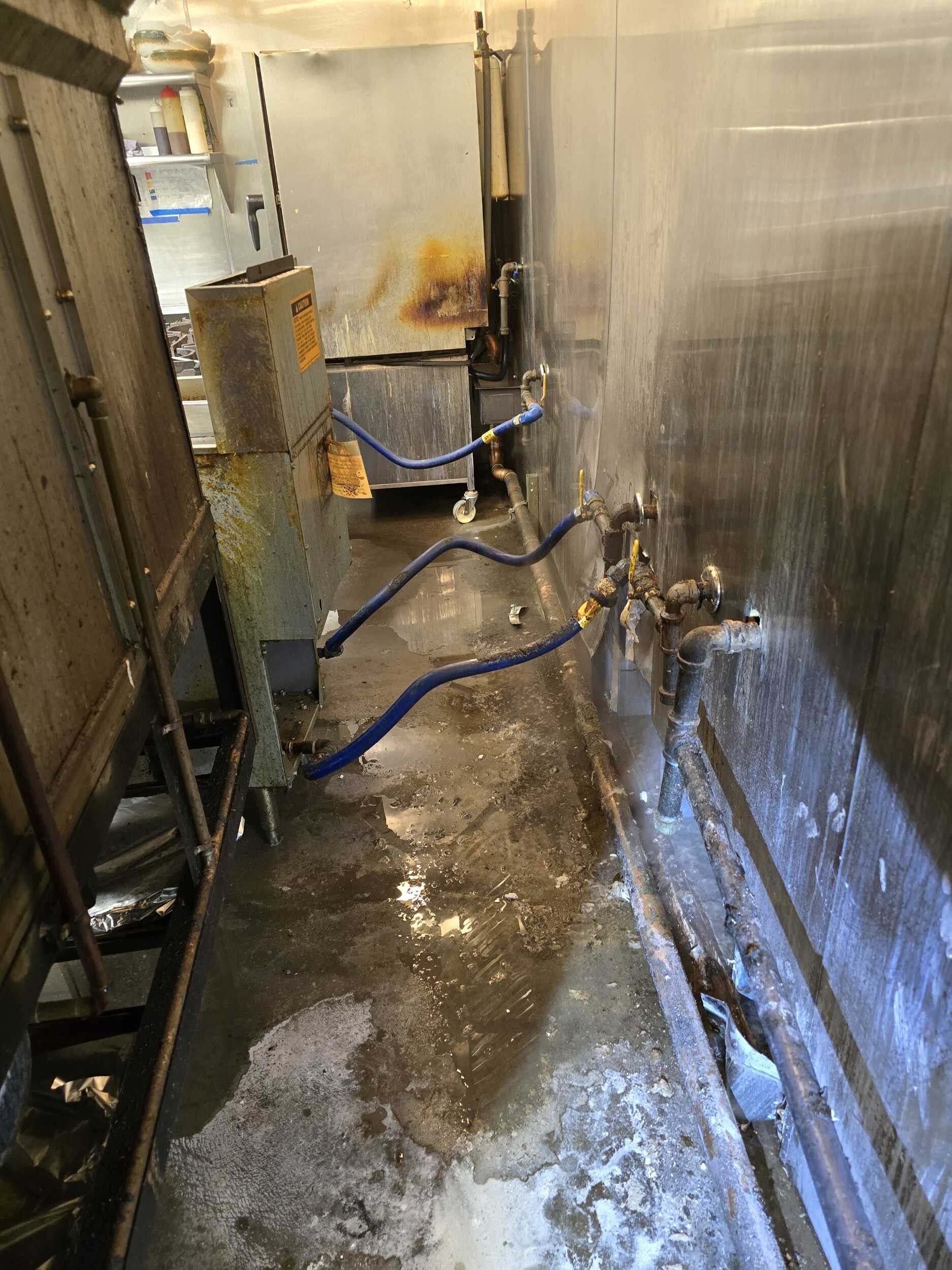Narrow industrial corridor with wet floor, blue hoses, stainless steel equipment, and metal walls.