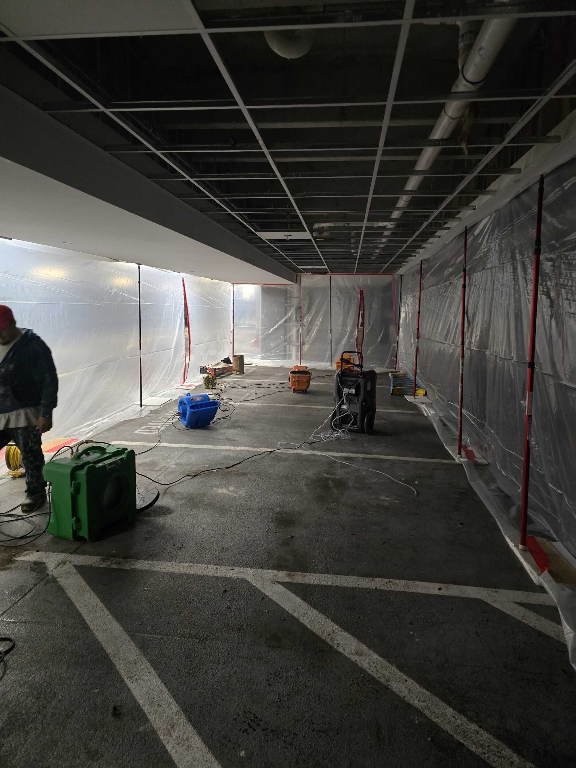Covered walkway or construction corridor with plastic sheeting, exposed ceiling, and equipment on the floor