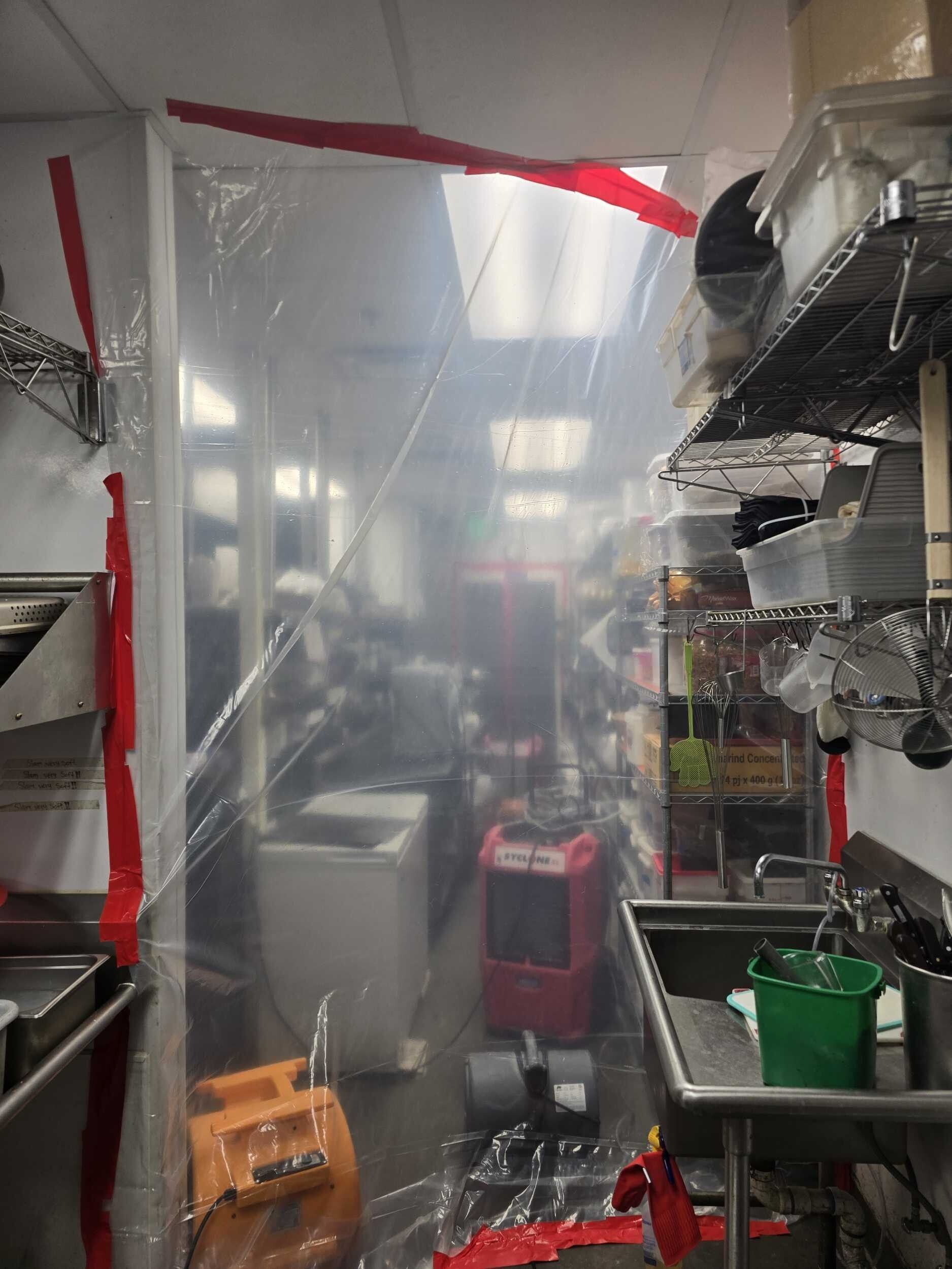 Narrow industrial kitchen with stainless shelves, clear plastic curtain, sink, and cleaning supplies.