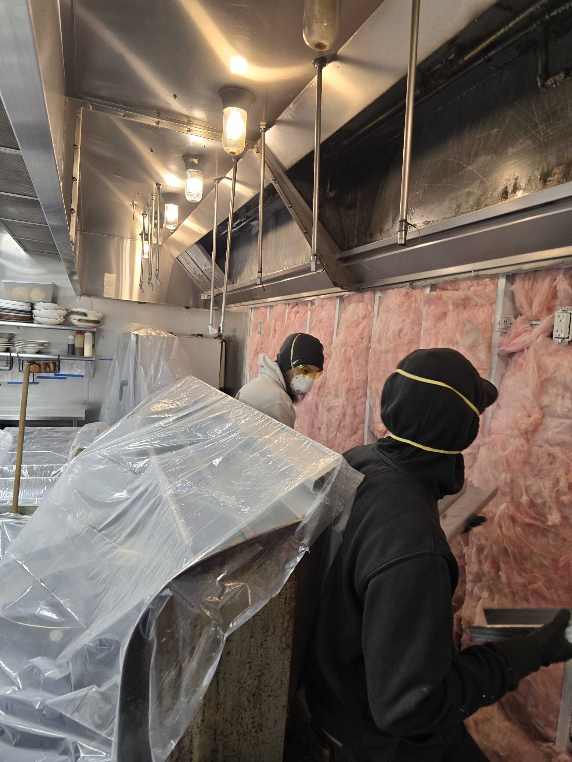 Worker installing pink insulation in a narrow attic space with exposed ducts and lights.