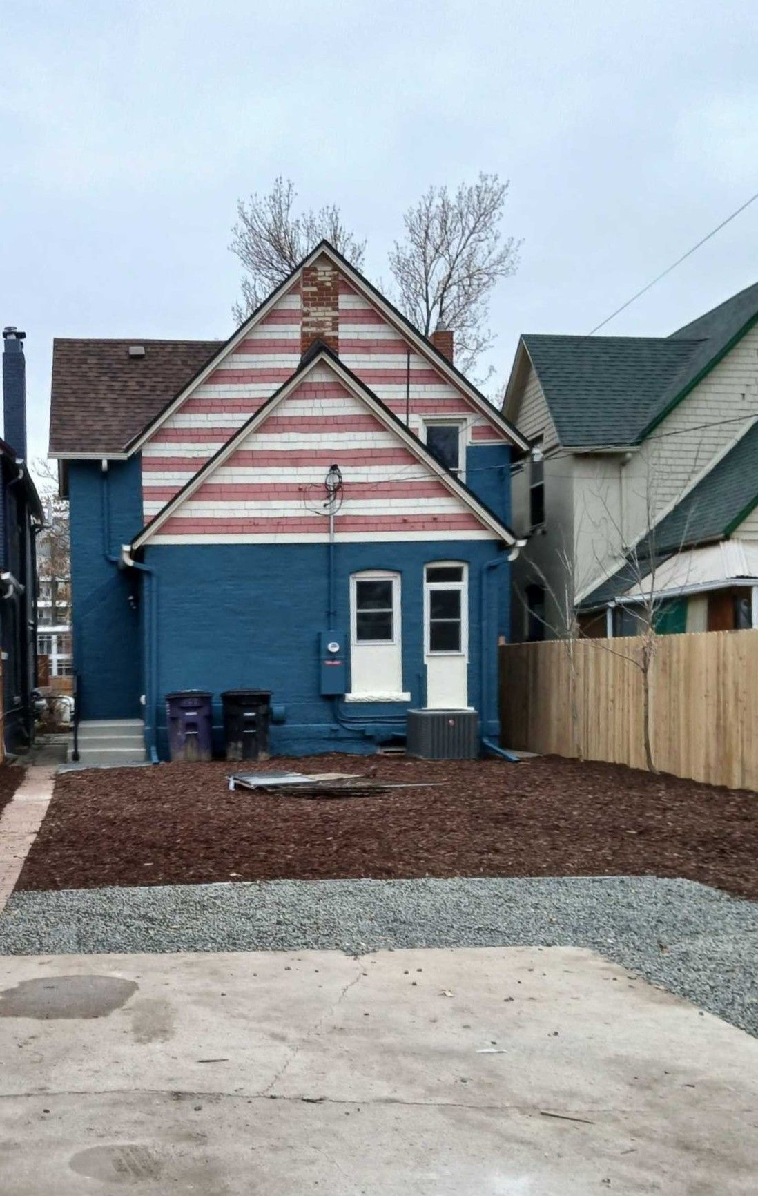Small blue house with pink-striped gable, gravel front yard, and neighboring homes under an overcast sky