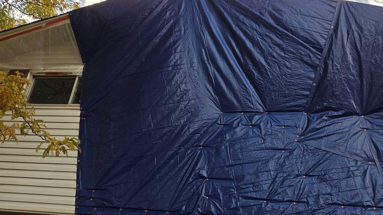 Blue tarp covering part of a house exterior beside a window and roofline