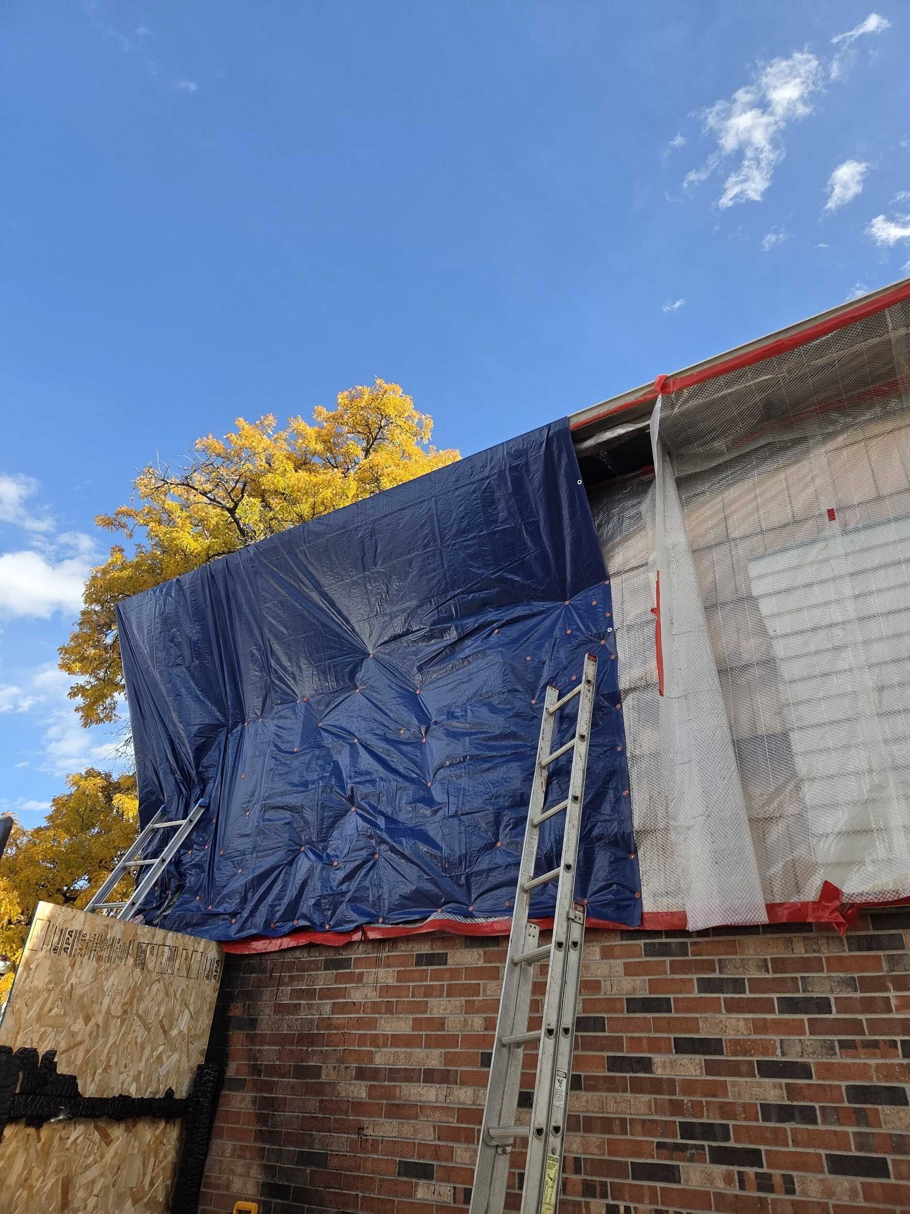 Scaffolding covered with blue tarp beside a brick wall and ladder under a blue sky