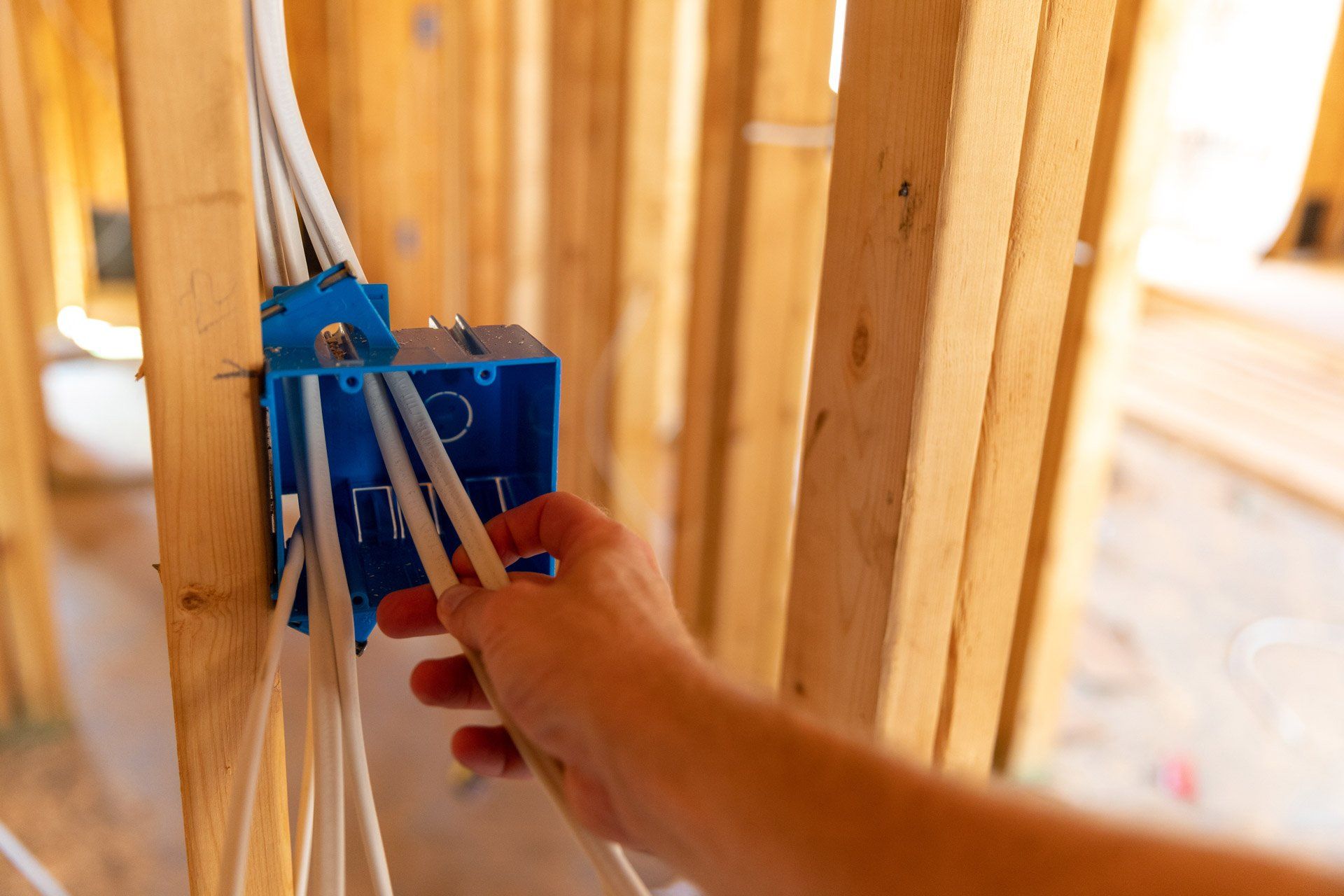 Hand pulling wires from a blue electrical box secured to wooden studs in a construction setting.
