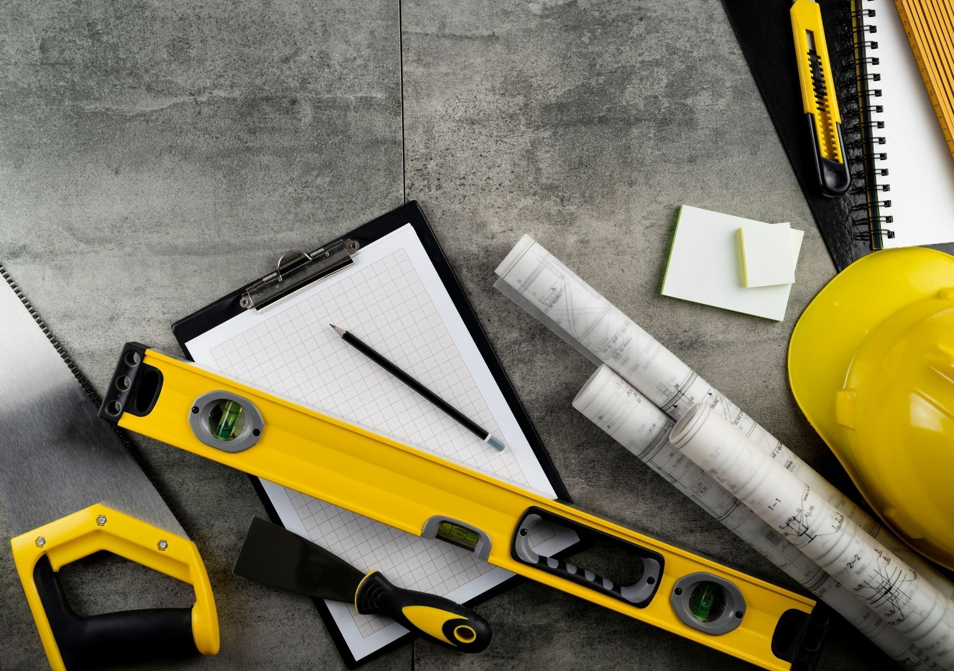 Construction tools on a gray surface: yellow hard hat, level, ruler, and notebook with pencil