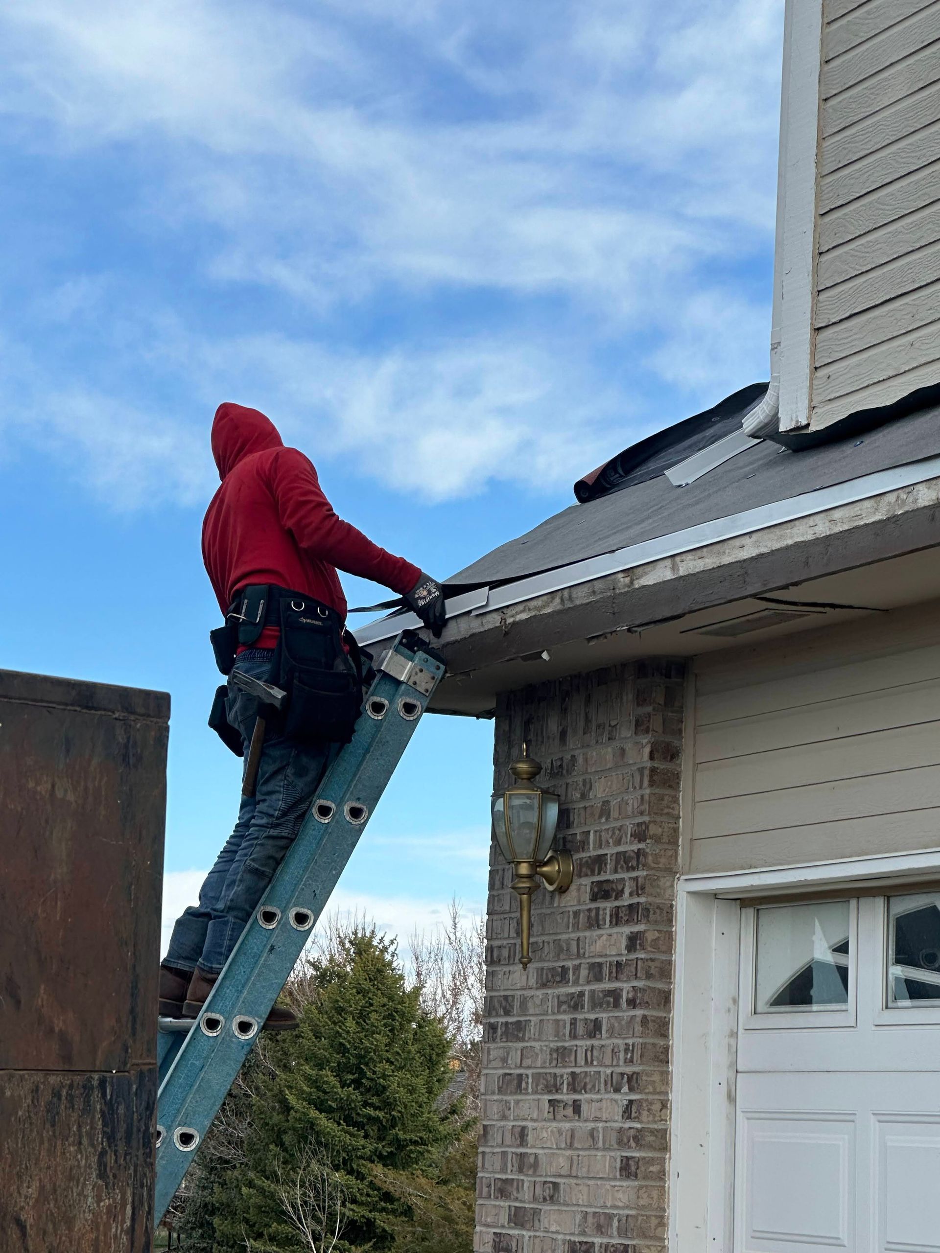 Worker on a ladder repairing a roof gutter beside a brick house under a blue sky